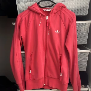 Rosa hoodie från Adidas med dragkedja - Säljer en snygg rosa hoodie från Adidas med klassiska tre ränder på ärmarna och vit logga på bröstet. Tröjan har huva med snörning, hel dragkedja och två fickor med dragkedja. Perfekt för en sportig eller lite yk ele 2000 look kanske