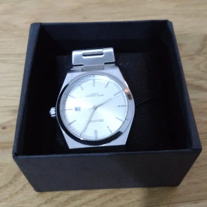 Silverfärgad klocka från Denvosi - Stilren klocka från Denvosi med silverfärgat armband i metall och vit urtavla med diskret mönster. Klockan har datumvisning och klassisk länkdesign. Perfekt accessoar för dig som gillar en clean och modern look.