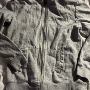 Grå zip hoodie från H&M BASIC - Enkel och stilren grå hoodie från H&M BASIC med dragkedja framtill och två fickor. Tillverkad i mjuk bomullsblandning, perfekt för chill dagar. Långärmad modell med ribbade muddar och klassisk passform.