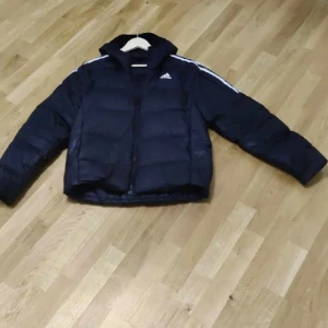 Svart pufferjacka från Adidas - Snygg svart pufferjacka från Adidas med klassiska vita ränder längs axlarna och Adidas-logga på bröstet. Jackan har huva, dragkedja framtill och är vadderad för extra värme. Perfekt för kalla dagar och streetwear-stil.