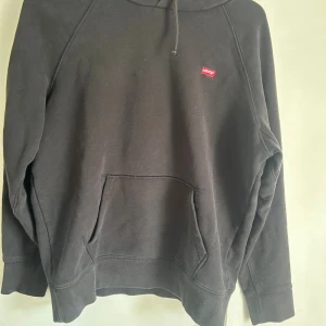 Svart hoodie från Levi's med logga - Svart hoodie från Levi's med klassisk röd logga på bröstet. Tröjan har huva med snörning och en stor magficka framtill. Perfekt för dig som gillar en enkel och stilren look. Mjuk och skön i bomullsmix.