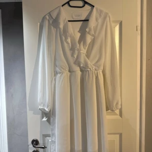 Vit långklänning med volang från Sisters Point - Lång, vit klänning från Sisters Point med v-ringning och volangdetaljer framtill. Klänningen har långa, transparenta ärmar med resår vid handlederna och markerad midja. Perfekt för dig som konfirmant eller student.