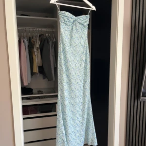Ljusblå långklänning från Nelly - Supersöt långklänning från Nelly i ljusblått med vitt och grönt blommigt mönster. Klänningen är axelbandslös med en snygg twist-detalj vid bysten. Den har använts en gång och har absolut inga defekter! Pris kan diskuteras.