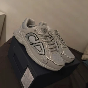 Dior B30 grå/vita sneakers stl 44 - Säljer ett par Dior B30 sneakers i grått och vitt med stora CD-loggor på sidorna. Skorna har mesh och mikrofiber i ovandelen, rund tå och snörning. Snygg chunky sula och stilren design som sticker ut. Perfekta för dig som gillar exklusiva och trendiga sneakers.
