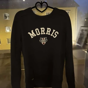 Svart tröja från Morris Gents XS - Svart långärmad tröja från Morris Gents i storlek XS. Tröjan har vit broderad logga och text framtill samt ribbade muddar vid ärmslut och nederkant. Klassisk och stilren modell som passar perfekt till jeans eller chinos. Sitter som S