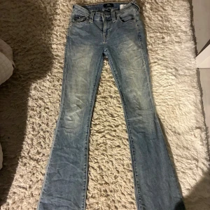 True Religion Becca Bootcut Jeans - Snygga ljusblå bootcut jeans från True Religion, modell Becca Mid Rise Bootcut. Jeansen har klassiska fickor med dekorativa sömmar och knappar bak, samt en bekväm midwaist-passform. Tillverkade i slitstarkt jeansmaterial och perfekta för en cool vardagslook.