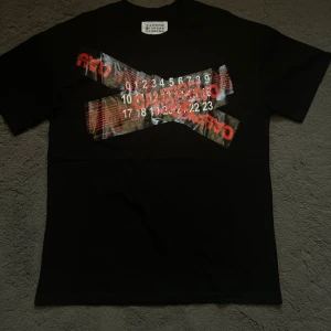 Svart t-shirt Maison Margiela - Svart t-shirt från Maison Margiela med ikonisk sifferetikett i nacken och coolt grafiskt tryck i rött och vitt framtill. Klassisk passform och rund halsringning. Perfekt för dig som gillar streetstyle och unika designer.