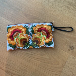 Broderad blommig kuvertväska - Färgstark kuvertväska med stora broderade blommor i gult, orange, grönt och rött på vit botten. Väskan har dragkedja upptill och en svart handledsrem. Vet tyvärr inte märket då den är köpt i Thailand❤️