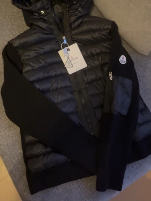 Svart Moncler Cardigan - Snygg svart Moncler Cardigan med stickade ärmar. Jackan har huva, dragkedja och en liten ficka med Moncler-logga på ärmen. Perfekt för kyliga dagar och riktigt trendig stil. Storlek L men passar S/M. Säljs då den inte passar mig och den är helt oanvänd.