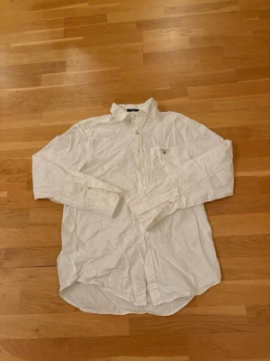 Vit oxfordskjorta från GANT - Klassisk vit oxfordskjorta från GANT i bomull med regular fit. Skjortan har lång ärm, knappar framtill och en bröstficka med liten broderad logga. Perfekt för en clean och stilren look.