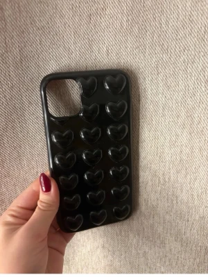 Svart mobilskal med hjärtan - Snyggt svart mobilskal med upphöjda hjärtan på baksidan. Skalets design är lekfull och trendig, perfekt för dig som vill ha något unikt till din mobil. Tillverkat i tåligt plastmaterial och passar modeller med större kameralins. Till iPhone 11