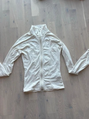 Vit ziptröja med hög krage - Säljer en stilren vit ziptröja med hög krage och långa ärmar. Tröjan har figurnära passform och dragkedja framtill. Perfekt att slänga på sig över en t-shirt. Materialet känns mjukt och lätt.