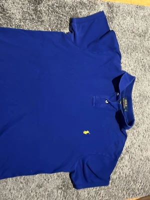 Blå pikétröja från Ralph Lauren - Klassisk blå pikétröja från Ralph Lauren med gul broderad logga på bröstet. Tröjan har krage, korta ärmar och knäppning vid halsen. Perfekt för en stilren och avslappnad look.