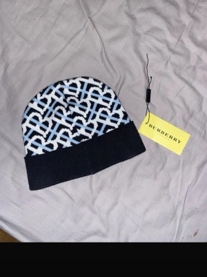 Burberry mönstrad mössa i blått och vitt - Snygg mössa från Burberry med ikoniskt monogrammönster i blått, vitt och svart. Mössan har en bred svart uppvikt kant och är stickad i mjukt material. Perfekt accessoar för att lyfta din outfit med en lyxig touch. Ge pris förslag