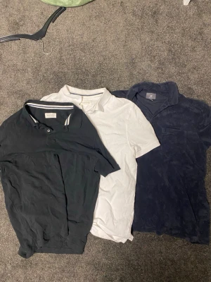 Tre pikétröjor i olika färger - Säljer tre pikétröjor i svart, vitt och mörkblått. Alla har klassisk krage och kort ärm, en av dem är från H&M och har frottéliknande material. Perfekta för en avslappnad stil. Alla är i L