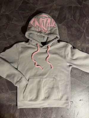 SYNA WORLD  - Snygg grå hoodie med huva och rosa snören. Huvan har ett coolt rosa graffiti-tryck framtill. Perfekt för dig som gillar streetwear och vill sticka ut lite extra. Stor känguruficka framtill och mjukt material.