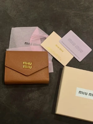 Brun plånbok från Miu Miu i läder - Snygg brun plånbok från Miu Miu i äkta läder med guldfärgad logga framtill. Plånboken har flera kortfack och ett fack för sedlar. Kommer med originalask och certifikat. Perfekt accessoar för dig som gillar stilrena detaljer.