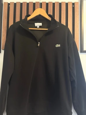 Svart halfzip tröja från Lacoste - Snygg svart tröja från Lacoste med klassisk halv dragkedja och det ikoniska krokodilmärket broderat på bröstet. Tröjan har en avslappnad passform och är tillverkad i mjukt bomullsmaterial. Perfekt för en stilren och enkel look.