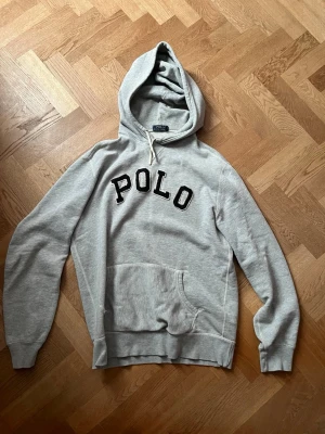 Grå hoodie från Polo Ralph Lauren - Snygg grå hoodie från Polo Ralph Lauren med stor svart POLO-text på bröstet. Klassisk känguruficka framtill, ribbade muddar och justerbar huva med dragsko. Tillverkad i mjuk bomullsblandning som är skön att ha på sig.