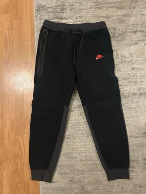 Svarta Nike Tech Fleece mjukisbyxor - Nike Tech Fleece mjukisbyxor i svart med grå detaljer längs benen och mudd vid benslut. Byxorna har dragsko i midjan, en dragkedjeförsedd ficka på sidan och rött Nike-logga tryckt på låret. Perfekta för en avslappnad och sportig stil.