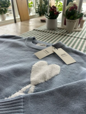 Ljusblå stickad tröja från Ami - Supersnygg ljusblå stickad tröja från Ami med ett stort vitt hjärta på bröstet. Tröjan har rund halsringning, ribbade muddar och nederkant. Perfekt för dig som vill ha en trendig och mysig look.