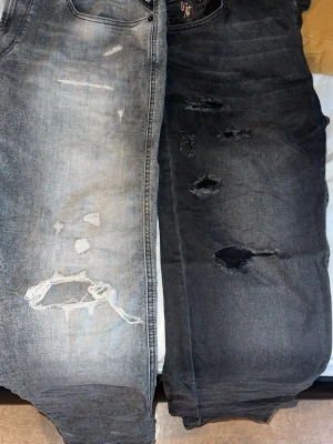 Två par slitna jeans, grå & svart - Säljer två par jeans, ett i ljusgrå tvätt och ett i svart, båda med slitningar och hål för en trendig look. Raka ben och klassisk femficksmodell. Perfekta för dig som gillar streetstyle och avslappnad vibe.