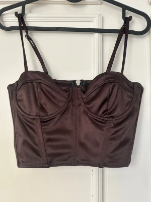 Brun bustier med smala axelband - Snygg brun bustier med smala justerbara axelband och markerade kupor. Toppen har en dragkedja bak och är sydd i ett glansigt, stretchigt material som ger en lyxig känsla. Perfekt för att skapa en trendig och självsäker look.