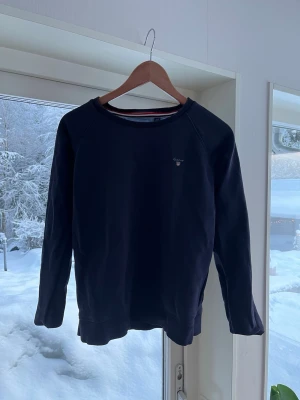 Mörkblå sweatshirt från Gant - Klassisk mörkblå sweatshirt från Gant med rund halsringning och diskret logobrodyr på bröstet. Tröjan har ribbade muddar vid ärmslut och nederkant samt en mjuk bomullskänsla. Perfekt för en avslappnad och stilren look.