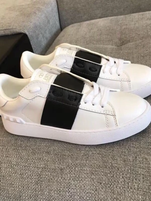 Vita sneakers med svart band Valentino - Snygga vita sneakers från Valentino med ett brett svart band över snörningen. Skorna har klassisk låg profil, vita snören och detaljer i vitt läder. Perfekta för dig som gillar stilrena och trendiga sneakers med en twist.