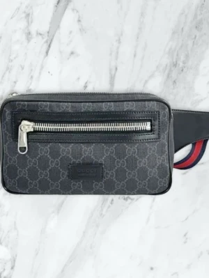 Gucci svart magväska med monogram - Snygg svart magväska från Gucci med klassiskt GG-monogram i grått. Väskan har ett stort fack med dragkedja framtill och justerbart band i rött och marinblått. Tillverkad i canvas med läderdetaljer och silverfärgad dragkedja. Perfekt accessoar för att hålla koll på dina grejer.