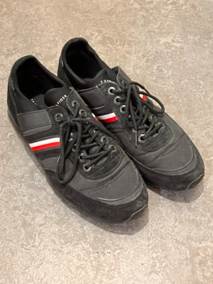 Svarta Tommy Hilfiger sneakers - Snygga svarta sneakers från Tommy Hilfiger med klassiska röda och vita ränder på sidan. Skorna har snörning, lågt skaft och är tillverkade i en mix av mocka och textil. Perfekta för dig som gillar stilrena och sportiga skor med en ikonisk touch.