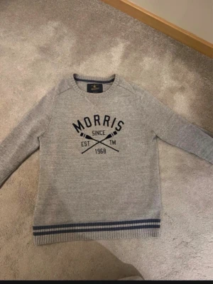 Grå tröja från Morris med tryck - Snygg grå långärmad tröja från Morris med svart tryck framtill och ribbade muddar. Tröjan har rund halsringning och blåa ränder längst ner vid midjan. Perfekt för en avslappnad och stilren look.