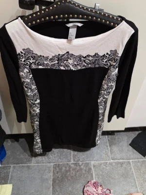 Svart och vit klänning från H&M - Snygg svart och vit klänning från H&M med 3/4-ärmar. Klänningen har ett blommigt spetsmönster upptill och längs sidorna, samt en kontrasterande svart mittdel. Rund halsringning och figurnära passform. Perfekt för dig som gillar stilrena detaljer.