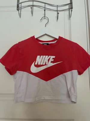 Nike croppad t-shirt röd och vit - Snygg croppad t-shirt från Nike i rött och vitt med stort Nike-logga framtill. T-shirten har korta ärmar och är tillverkad i mjuk bomull, som ny. Knappt använd 