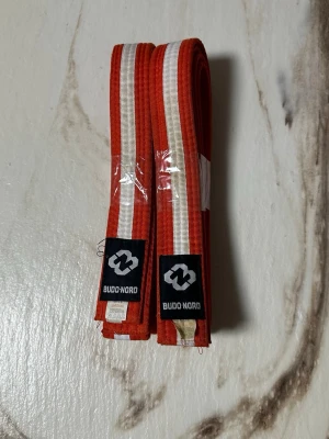 Rödvit bältespar från Budo-Nord - Två stycken rödvita bälten från Budo-Nord, perfekta för kampsport som judo eller karate. Bältena är tillverkade i bomull och har en tydlig vit rand i mitten. Märkesetikett sitter fastsydd på ena änden.
