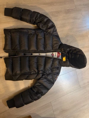 Svart pufferjacka Parajumpers Sheen - Svart pufferjacka från Parajumpers, modell Sheen. Jackan har en vadderad design, huva och dragkedja framtill som tyvärr har gått sönder, dock är den fortfarande varm så funkar ju om man inte vill stänga den. Detaljer med gul rem vid halsen och ribbade muddar vid ärmslut. Perfekt för kalla vinterdagar och riktigt snygg streetstil.