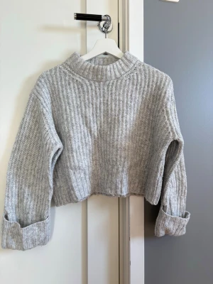 Grå croppad stickad tröja från Gina Tricot - Säljer en grå croppad stickad tröja med bred ribbstickning och uppvikta ärmslut. Tröjan har en hög, ribbad krage.