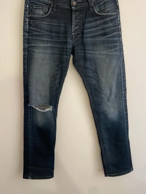 Mörkblå jeans från Jack and Jones  - Säljer ett par mörkblå jeans med tvättade detaljer och ett snyggt hål på vänster knä. Jeansen har fem fickor, raka ben och klassisk knappgylf. Materialet är denim i bomull och passformen är smal. Perfekta för en avslappnad och trendig look.