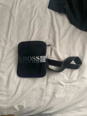 Svart axelväska från Hugo Boss - Snygg svart axelväska från Hugo Boss med justerbar axelrem och logga i vitt framtill. Väskan har en fyrkantig form och dragkedja upptill. Perfekt för att bära det viktigaste och ger en clean streetwear-look.