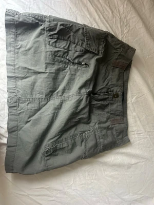 Grå cargokjol med fickor - Skitsnygg grå cargokjol. Kjolen har en kort passform och stängs med knapp och dragkedja framtill. Perfekt för en avslappnad och cool stil. Använd väldigt få gånger. 