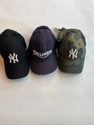 Camo New Era NY keps Triumph - Snygg keps från New Era med klassiskt NY-broderi framtill. Kepsen har ett camouflagemönster i gröna och bruna toner samt böjd skärm. Perfekt accessoar för en streetwear-look. Och en två andra en triumph och en svart