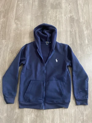 Mörkblå hoodie från Polo Ralph Lauren - Snygg mörkblå hoodie med dragkedja från Polo Ralph Lauren. Klassisk design med vit broderad logga på bröstet, två fickor fram och skön huva. Perfekt för en avslappnad och stilren look.