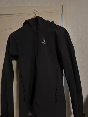 Haglöfs vindjacka  - Snygg svart vindjacka från Haglöfs med huva och hel dragkedja framtill. Jackan har två snedställda fickor med dragkedja och diskret Haglöfs-logga på bröstet. Perfekt för blåsiga dagar och enkel att matcha med olika outfits.