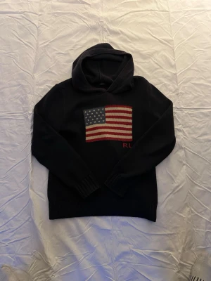 Svart hoodie från Ralph Lauren - Snygg svart hoodie från Ralph Lauren med stickad amerikansk flagga på bröstet och RL-broderi under. Hoodien har ribbade muddar vid ärmslut och nederkant samt en klassisk huva. Perfekt för dig som vill ha en stilren och ikonisk look.