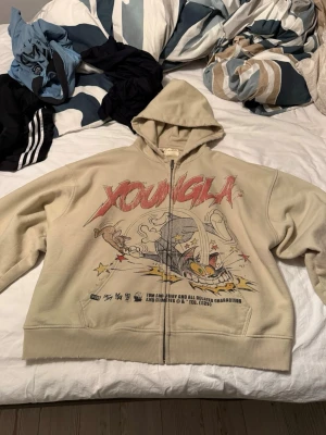 Beige Tom & Jerry hoodie från Youngla - Snygg beige hoodie från Youngla med dragkedja och stor Tom & Jerry-tryck på framsidan. Använd max 5 gånger säljer pga att den är för kort för mig. Helt slut på younglas hemsida