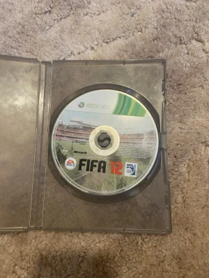 FIFA 12 till Xbox 360 - FIFA 12 till Xbox 360, ett klassiskt fotbollsspel från EA Sports. Skivan är rund med grön och vit design samt FIFA-logga. Perfekt för dig som gillar sportspel och vill spela med vänner på konsol.