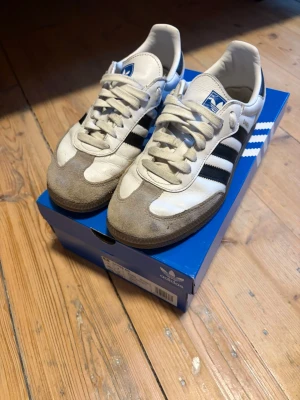 Adidas Samba vita och svarta sneakers - Klassiska Adidas Samba sneakers i vitt läder med svarta ränder och grå mockadetaljer på tån. Låg modell med vit sula och blå logga på plösen. Perfekta för en sportig och retro stil.