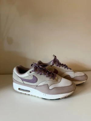 Nike Air Max 1 beige/lila sneakers - Nike Air Max 1 sneakers i beige och lila med klassisk siluett. Ovandel i mesh och mocka, lila swoosh och matchande skosnören. Vit mellansula med synlig Air-enhet och beige detaljer. Perfekta för dig som gillar retrostil och snygga färgkombos.