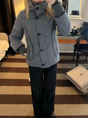 Grå vinterjacka med knappar - Snygg grå jacka med hög krage och stora knappar framtill. Jackan har långa ärmar, två fickor och smockdetaljer i midjan för en schysst passform. Perfekt för kyligare dagar och enkel att matcha med olika outfits.