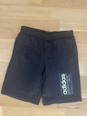 Svarta shorts från adidas med tryck - Svarta shorts från adidas med vitt tryck på ena benet och bred resår i midjan. Klassisk sportig look med snörning i midjan för bästa passform. Perfekta för träning eller chill dagar.